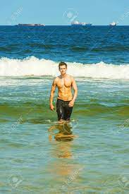 Portrait Of Young Sexy Man. Well Built Young Handsome Muscular Guy, Half  Naked, Wearing Bathing Suit, Walking Out Of Water On The Beach, Narrowing  Eyes Under Strong Sun Shine Stock Photo, Picture