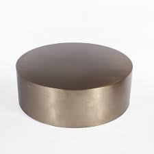 This contemporary piece sits on a circular metal drum base with a gold finish and lower shelf. Fedora Coffee Table In Brass Mid Mod