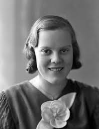 Foto av en kvinna i klänning med en tygblomma mitt fram. Bröstbild, en  face. Ateljéfoto. 'Målare