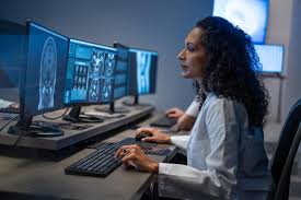 A professional consultation setting with a medical professional sitting at a desk facing a client. The room has a modern aesthetic with white walls decorated with framed certificates. The desk is organized with office supplies, a laptop, and a fruit bowl in the center.
