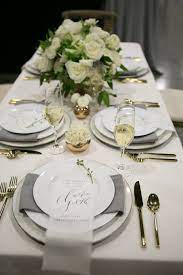 The setting consists of a service plate in the center, with forks set to the left of the service plate and knives and spoons set to the right. Elegant White Table Setting By Stem Floral Design Dinner Table Decor Dinner Table Setting Dinner Party Table