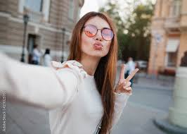 Selfie-portrait of pretty girl with long brunette hair at street  background. wears beige dress, red sunglasses, make up. Smiling and  positive.young woman in glasses making selfie , showing two fingers Stock  Photo |
