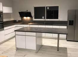 Aménagement d'une cuisine ouverte contemporaine en l avec un placard à porte plane, des portes de placard en bois sombre, un plan de travail en inox, une crédence blanche, un électroménager blanc, un îlot central, un sol gris et un plan de travail gris. Quel Plan De Travail Choisir Pour Une Cuisine Blanche Comera Cuisines