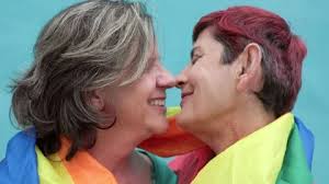 Happy gay senior lesbian couple kissing wearing lgbt rainbow flag outdoors