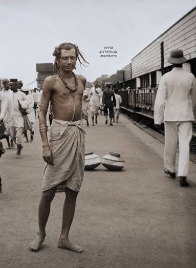 Passengers on the Great Indian Peninsula Railway (G.I.P. Railway), July 1906. An ascetic (sadhu) stands on a busy railway platform as passengers go about their journeys. Restored and colorized from an original