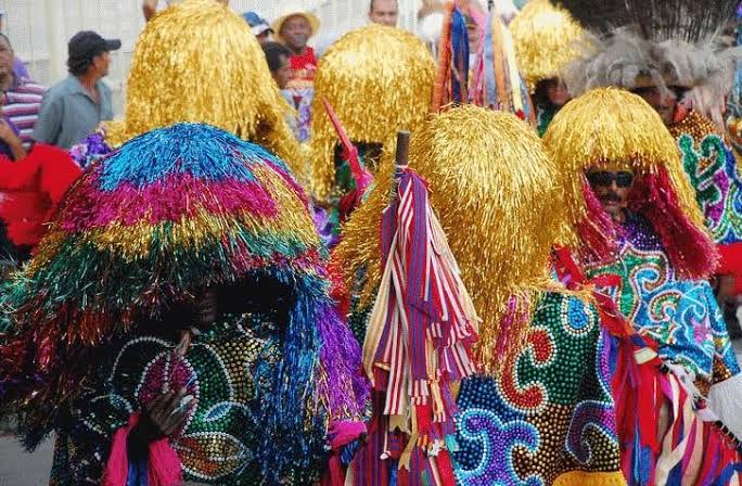 Grupo de pessoas participa de uma manifestação cultural popular ao ar livre. Os participantes usam fantasias coloridas com roupas bordadas por lantejoulas e desenhos em espiral, além de grandes máscaras ou capacetes cobertos por longas franjas metálicas douradas e multicoloridas, que escondem completamente o rosto. Um dos participantes à direita usa óculos escuros visíveis sob a franja. Ao centro, há um estandarte decorado com fitas coloridas. Ao fundo, outras pessoas observam o evento, reforçando o clima de festa tradicional.