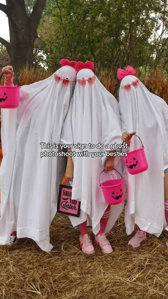 Three people dressed as classic white sheet ghosts styled with hot pink hair bows, heart-shaped sunglasses, and holding pink pumpkin buckets.