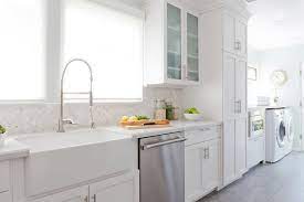 Painted in benjamin moore hale navy, navy blue shaker cabinets, accented with white and gray quartzite countertops, flank a wolf range sat under a navy blue hood framed with a marble herringbone backsplash and positioned between windows dressed in bamboo roman shades illuminated by swing arm brass sconces. Kitchen And Laundry Room Combo Design Transitional Kitchen Kitchen Kitchen Remodel Long Kitchen
