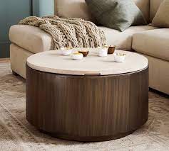 A large storage ottoman with a hidden compartment underneath the top lid, made out of faux leather in a black color padded around the solid wood structure and paired with an additional wooden top to make it stable. Gilman 30 Round Storage Coffee Table Pottery Barn