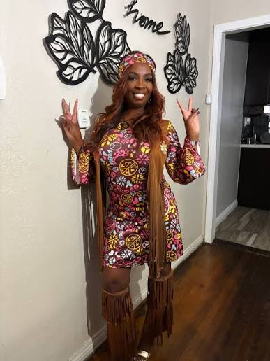 Smiling woman posing in a 1960s hippie costume with a colorful peace-sign print dress, long brown fringe vest, matching headband, and fringe leg covers while holding up peace signs.
