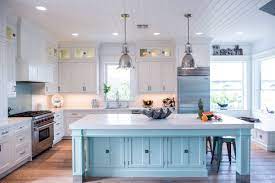 A gorgeous coastal kitchen with light blue cabinets, a light blue kitchen island, rattan stools and woven pendant lamps, potted greenery. Coastal Style White Kitchen With Blue Island Crystal Cabinets
