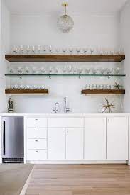 These floating shelves will not take much space on the wall will be a mystery themselves. A Smoke Gray Glass Globe Light Lights A White Wet Bar Boasting Stacked Brown Wood And Glass Floating Shelves Glass Shelves Glass Bar Shelves Glass Wall Shelves