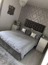 Bedroom features a gray print bed with white bedding and purple and gray pillows flanked by cream lattice bedside tables lit by gray glass lamps on white plank walls, purple stools at the foot of the bed, a gray corner chair, and a white lamp on a white dresser, lit by white shaded hanging lamps. Beautiful Grey Bedroom Ideas With Mirrored Furniture To Inspire You Schlafzimmer Design Hauptschlafzimmer Wohnung Schlafzimmer