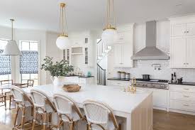 Contemporary arrangement for a spacious kitchen, furnished with a rectangular kitchen island in the middle with an additional bar with a marble top surface, paired with three barstools. Welcoming White Kitchen Boasts Serena Lily Riviera Counter Stools Placed In Front Of An Off Bistro Kitchen Stools For Kitchen Island White Marble Countertops
