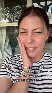 Lauren has brown hair tied back in a pony tail. She is wearing a black and  white striped t shirt. Lauren is in front of a green home and points to  black and green Halloween pumpkins.