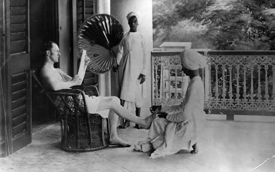 A British officer reading a newspaper while being fanned with a palm frond & getting a pedicure from one of his servants in India, late 1800's