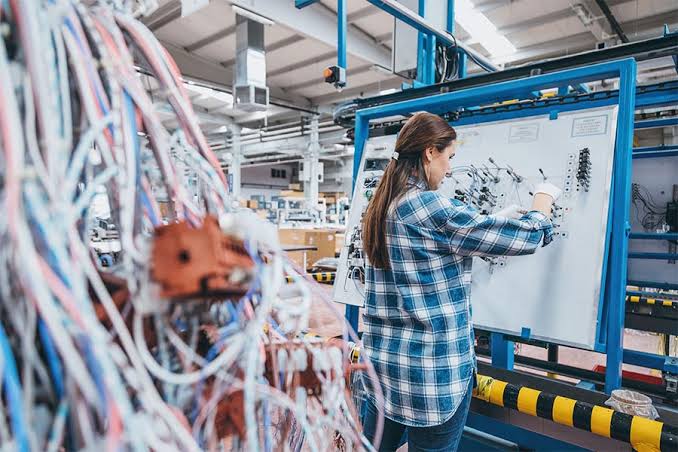 Technicians assembling wiring harnesses on a factory production line, cutting, crimping, and connecting cables at organized workstations under bright industrial lighting.