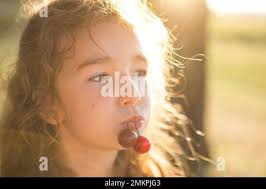 A cheerful sweet girl with cherry berries in her mouth. Funny summer  portrait of a child with a cherry, gifts of summer, summer time Stock Photo 