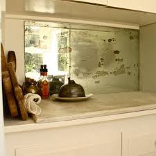 A white and gray marble countertop flanks a wolf range placed against mirrored backsplash tiles beneath a stainless steel hood accented with brass. Smokey Mirrored Splashback Antique Mirror Backsplash Mirror Backsplash Kitchen Mirror