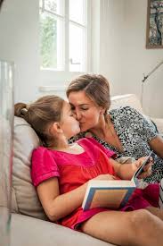 Mother kissing daughter with book on couch in living room stock photo