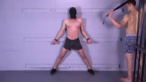 Logan is bound to the wall, hood removed and whipped by sexy muscled captor