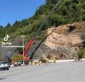 A backhoe resting on a platform suspended by a crane, with ropes attached to corners for control by two guys on the ground, excavating a hill?!