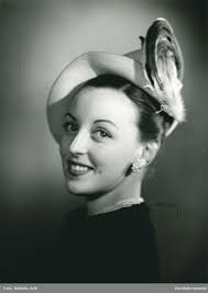 Porträtt av kvinna i hatt av Suzanne Talbot. Gul stråfjäder med svart  sammetsgarnering, svart crasse, samt gult flor med svarta moucher.…