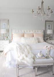 A bright feminine bedroom with a cnaopy bed, a wooden bench and a woven chair, a statement plant and a floral chandelier. Blush Pink Bedding Pom Pom At Home Romantic Homes Romantic Feminine Bedroom Faux Fur Throw Blanke Feminine Bedroom Master Bedroom Update Home Decor Bedroom