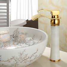 White and gold bathroom features a white washstand topped with white marble under a gold arched mirror lit by gold wall sconces and a small barbara barry simple scallop pendant. Gold Polished Brass Waterfall Bathroom Bow Sink Faucet Tall Single Hole White Marble Jade