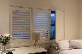 Taupe silk curtains cover windows fitted with white shutters and positioned behind a blue wingback headboard accenting a bed dressed in white bedding. Plantation Shutters Prices Plantation Blinds Timber Shutters Cost
