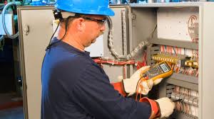 안전 장구를 착용한 엔지니어가 산업용 배전반 앞에서 노란색 멀티미터로 전압을 측정하는 현장 사진