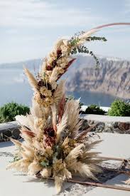 Find some very large foliage branches for this installation, as these very quickly build up the foam and coverage of the arch. Modern Bohemian Pampas Wedding In Greece Tie The Knot Santorini Dried Flowers Wedding Greece Wedding Dried Flowers