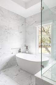 Marble tiled bathtub with a gold deck mount tub filler next to a walk in shower designed with marble floors and white marble trim. Marble Bathroom Niche Design Ideas