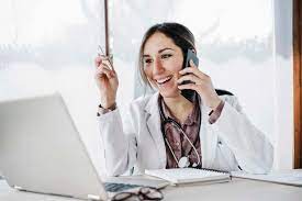 Lächelnd weiblichen Arzt Blick auf Laptop während des Gesprächs auf Handy  im Krankenhaus, lizenzfreies Stockfoto