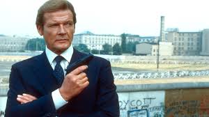 Immagine di Roger Moore nella veste di James Bond, in giacca scura, cravatta e camicia bianca, inappuntabile, braccia incrociate con una pistola sulla mano destra, sguardo cinico verso un punto lontano. Sullo sfondo l'immagine di una città