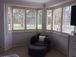 A white loveseat topped with blue pillows is positioned in front of windows covered in plantation shutters layered behind white curtains. Bay Window Shutters Specialists Australian Plantation Shutters