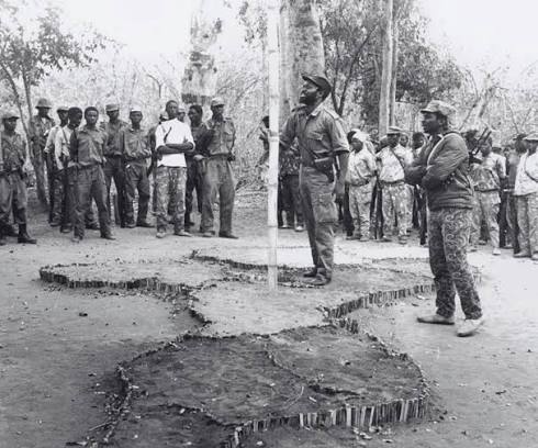 Deep inside a FRELIMO-controlled zone in Mozambique, Samora Machel  addresses guerrilla fighters, teachers and students in 1974. At the centre  of the improvised clearing, the future Mozambican leader stands beside a  hand-crafted