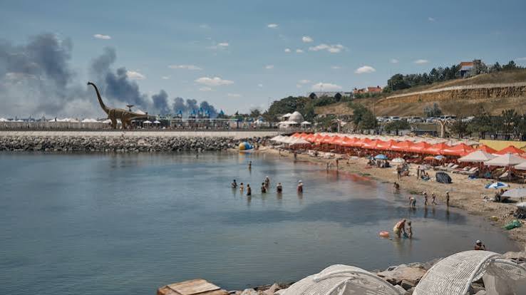 Cargo ships stalled in the Black Sea near the port of Odessa, with grain silos visible in the background under smoke.