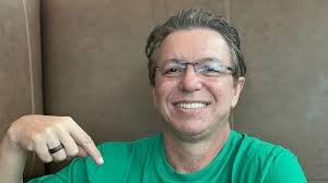 Homem sorri e aponta para baixo, usando óculos e camiseta verde.