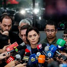 Mulher cercada por microfones de imprensa, com pessoas ao fundo. Possível coletiva de imprensa.