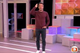 Tadeu Schmidt, apresentador do BBB, em um cenário moderno com luzes coloridas e formas geométricas.