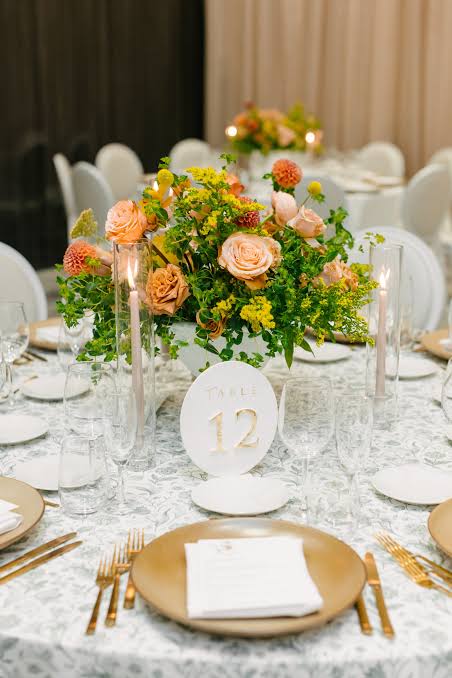 A round table set for an elegant event, featuring a centerpiece of orange  roses, yellow flowers,
