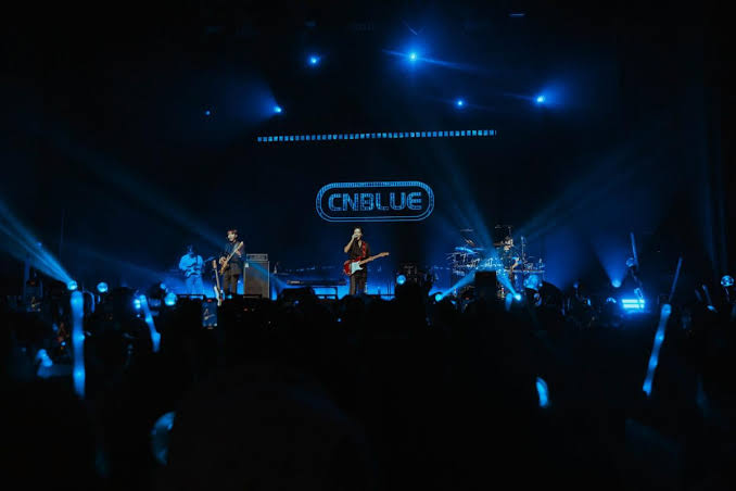 CNBLUE performs live on a dimly lit stage illuminated by vibrant blue spotlights, facing a large crowd holding glowing blue lightsticks.