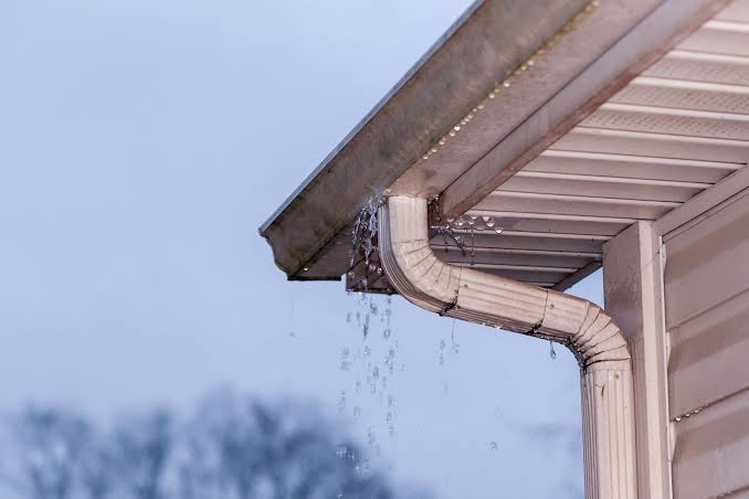 Close-up of a home gutter and downspout overflowing with rainwater during a storm, highlighting water drainage issues along the roofline.