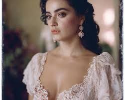 Dark-haired woman in vintage lace dress and earrings gazes intently with  blurred floral background