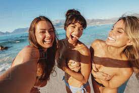 Three female friends taking selfie by the sea and laughing. Topless young  women enjoying summer on the beach. - Stock Photo - Dissolve