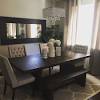 It has dark brown elegant dining table and chairs that go well with the intricate dark brown wallpaper that is augmented by various potted plants and flowers. 1