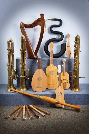 Conjunto de instrumentos musicais antigos expostos sobre um piso cinza, organizados em diferentes níveis. Ao fundo, há uma harpa de madeira, um grande instrumento de sopro curvo de cor preta e dois instrumentos de metal dourado semelhantes a trompetes ou serpentões, posicionados nas laterais. No centro, estão quatro instrumentos de cordas de madeira em tamanhos variados, semelhantes a alaúdes, violões antigos e uma pequena rabeca. Na parte inferior da imagem, há um instrumento de sopro longo de madeira, disposto na horizontal, e diversos instrumentos menores, também de madeira, alinhados lado a lado. O conjunto representa diferentes famílias de instrumentos musicais históricos, incluindo cordofones, aerofones e instrumentos de sopro de diferentes épocas.