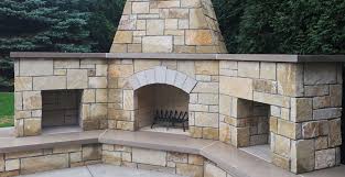 The fireplace is situated on a stone patio and accentuated with weaved wengue chairs and tables in striped black and white upholstery. Fireplace Stone Veneer Modern Rustic Outdoor Patio Dream Home Landscape Buechel Stone