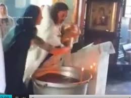 Un prêtre tient dans ses bras un nourrisson, devant trois proches qui l'observent à quelques centimètres de lui. Un Pretre Suspendu Apres Un Bapteme Violent Sur Un Bebe Video